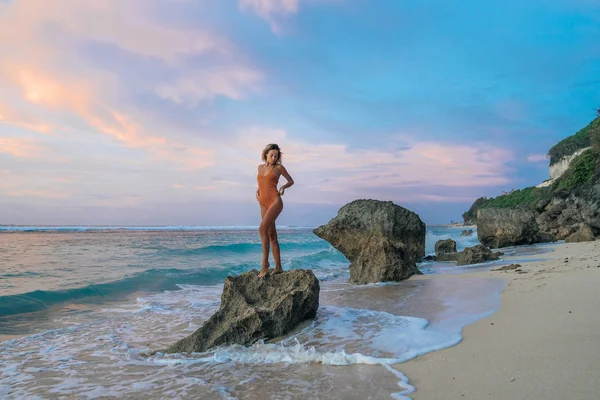 Slim meisje in badpak staande op een grote steen op wild beach achtergrond van prachtige zonsondergang — Stockfoto