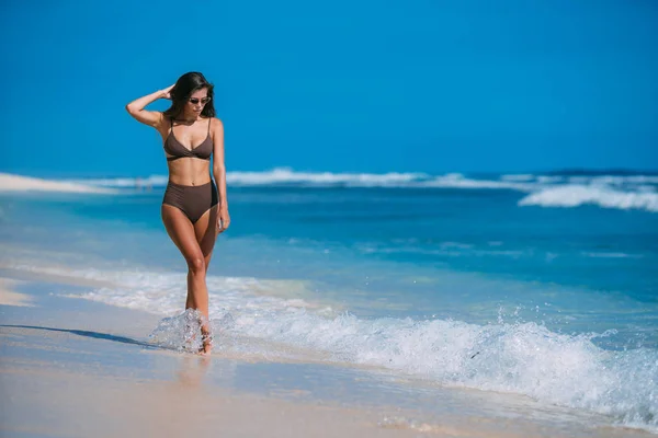 Modelo sexy em maiô marrom e óculos de sol posando na praia de areia branca — Fotografia de Stock