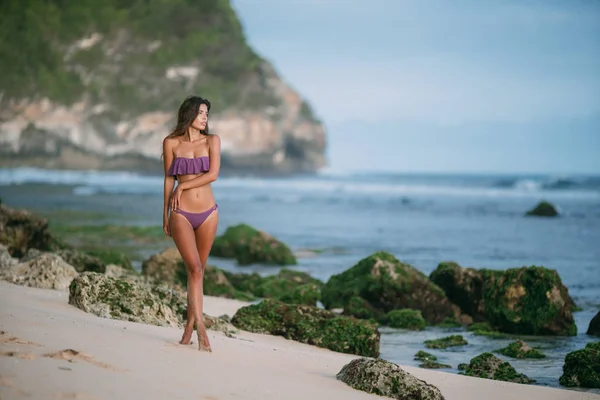 Chica bronceada sexy delgada en traje de baño posando en la playa con arena y piedras grandes —  Fotos de Stock