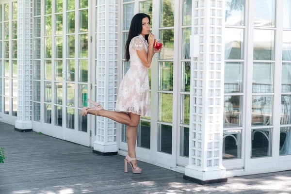 Belle fille brune avec verre de limonade posant en robe de couleur beige à l'extérieur d'un café près d'un mur en bois et en verre — Photo