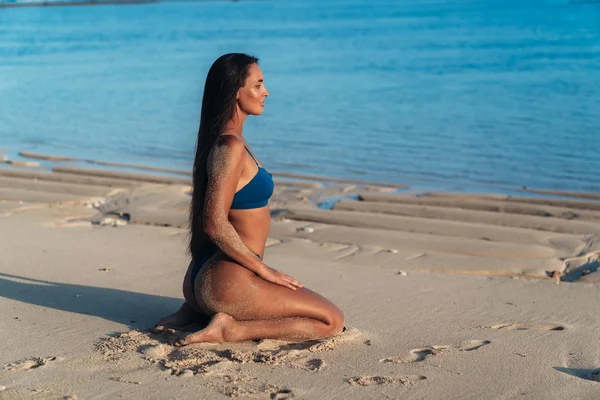 Schöne sexy Mädchen mit langen Haaren im blauen Bikini sitzt am Strand mit weißem Sand. — Stockfoto