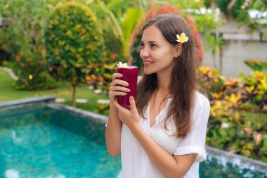 Tropikal bahçede çiçek saç ve bardak taze meyve suyu ile güzel kız duruyor.