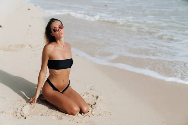 Чуттєва засмагла сексуальна дівчина в купальнику і сонцезахисних окулярах позує на пляжі з білим піском — стокове фото