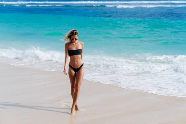 Abbronzata ragazza in costume da bagno nero e occhiali da sole passeggiate lungo la riva della spiaggia — Foto Stock