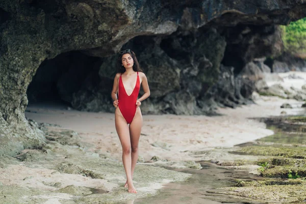 Sottile ragazza abbronzata in costumi da bagno e occhiali da sole in posa vicino alla pietra sulla spiaggia rocciosa dall'oceano . — Foto Stock