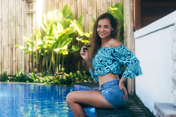 Chica sexy feliz en pantalones cortos de mezclilla y la parte superior se sienta en la piscina. Joven modelo bronceada descansando en el día de verano — Foto de Stock