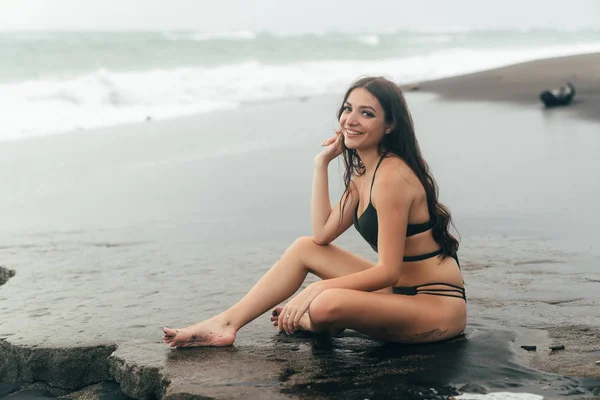 Vista lateral de chica sexy en bikini pasa tiempo en la playa de arena negra. Mujer joven con el pelo largo posando al aire libre —  Fotos de Stock