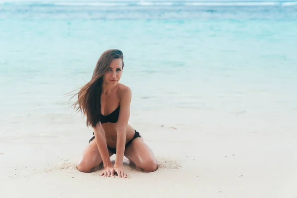 Σέξι μαυρισμένο κορίτσι στο μαύρο μαγιό ποζάρει στην αμμώδη παραλία κοντά στον ωκεανό. Το όμορφο μοντέλο sunbathes και στηρίζεται — Φωτογραφία Αρχείου