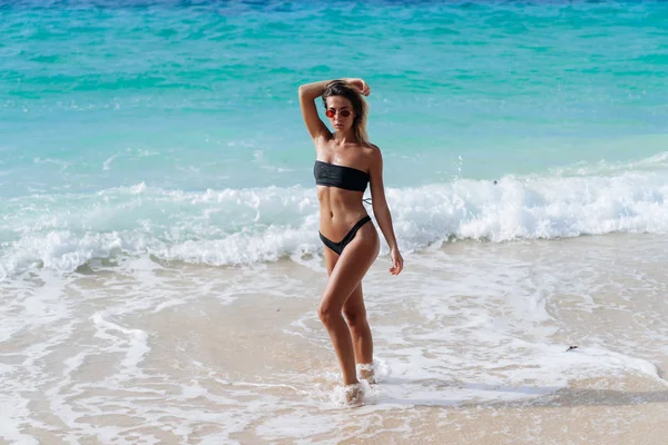 Bella ragazza in costume da bagno nero e occhiali da sole a riposo vicino all'oceano sulla spiaggia — Foto Stock