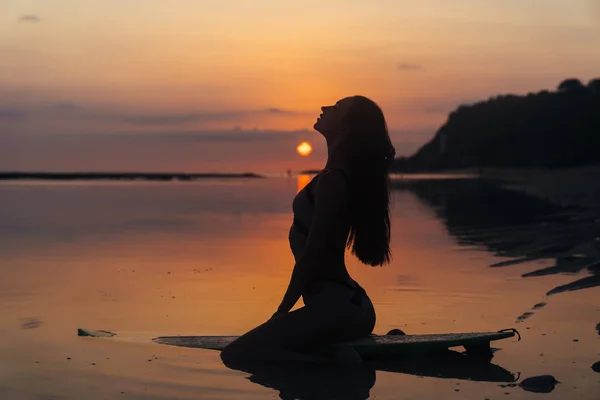 Silueta sexy dívka v plavkách lhaní a Pózování na surfovací prkno na pláži při západu slunce — Stock fotografie
