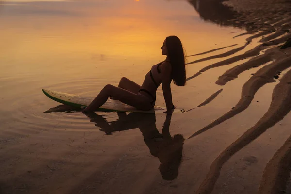 在美丽的日落背景下, 在海边的冲浪板上坐着的女孩的剪影和倒影 — 图库照片