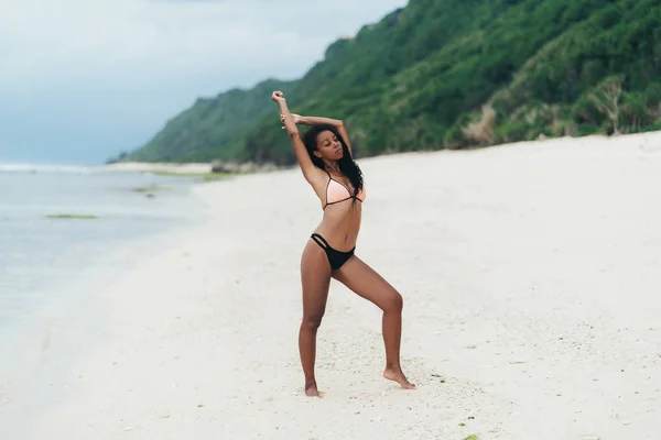 Sexy chica afroamericana en traje de baño descansando en la playa del océano. Joven mujer de piel negra con el pelo rizado se levanta en la orilla del mar —  Fotos de Stock
