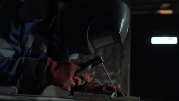 Slow-motion, side view welder in welding mask welds two metal parts. Workman in coveralls is working indoors — Stock Video