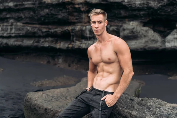 Kara kumlu plajda taş üzerinde siyah pantolonlu kaslı seksi adam