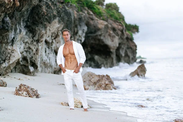 흰색 셔츠와 바지에 섹시 한 검게 그을린 남자가 해변에 쉬고, 배경에서 바다 파도. — 스톡 사진