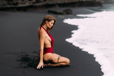 Young sexy girl with big breasts in a red swimsuit resting on black sand beach.