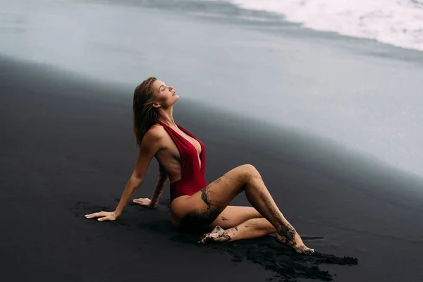 Junges sexy Mädchen mit großen Brüsten im roten Badeanzug am schwarzen Sandstrand. — Stockfoto