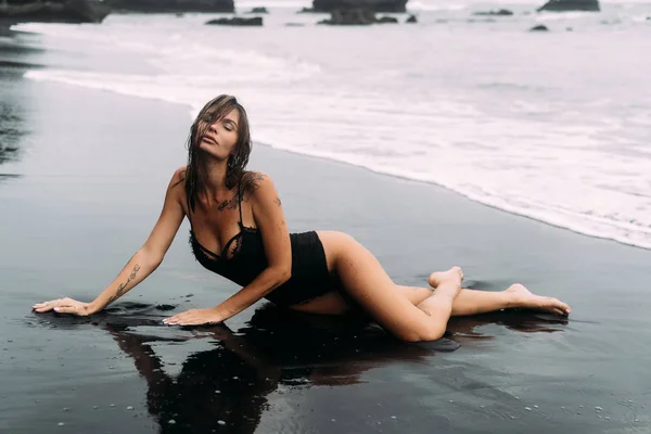 Menina loira sexy em roupa de banho preta repousa na praia de areia preta . — Fotografia de Stock