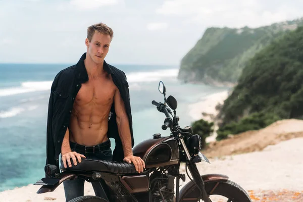 Portrait of sexy athletic man with naked torso near custom motorbike, ocean at background