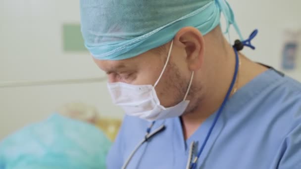 Retrato de cerca del médico en máscara estéril y gorra en la sala de cirugía durante la operación — Vídeo de stock