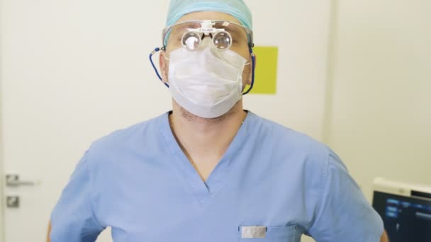 Ο γιατρός αφαιρεί τα μεγεθυντικά κιάλια και τη αποστειρωμένη μάσκα, κοιτάζει την κάμερα και χαμογελά — Αρχείο Βίντεο