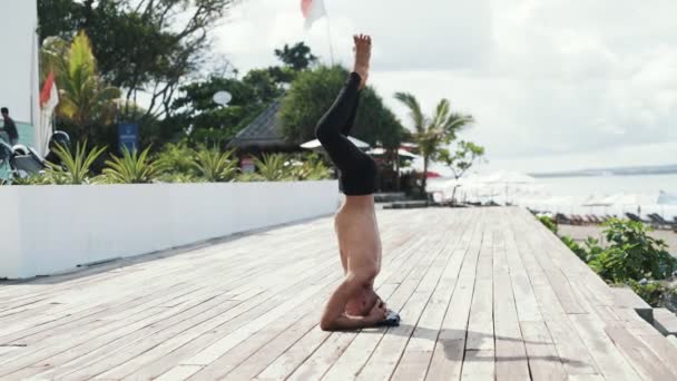 Hombre deportivo de cámara lenta haciendo ejercicio de yoga de pie sobre la cabeza, el mar y las palmas en el fondo — Vídeos de Stock