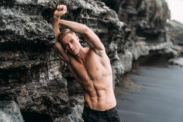 Sexy fitness male model in black pants and shirtless posing on black sandy beach.