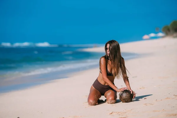 Modelo bronceado sexy con gafas de sol en traje de baño marrón con posando coco en la playa de arena blanca — Foto de Stock