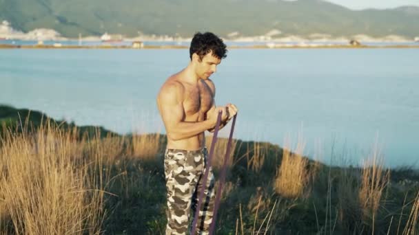 Atleta haciendo ejercicios con una banda elástica de fitness sobre un fondo de montañas y el mar . — Vídeos de Stock