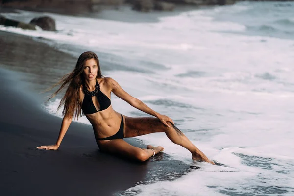 Young sexy girl in black swimwear resting on black volcanic sand beach. — Stock Photo, Image