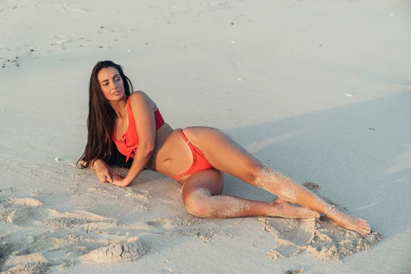 Retrato de menina bonita com cabelos longos em maiô vermelho deitado na praia com areia branca — Fotografia de Stock