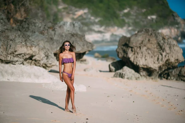 Sexy chica bronceada en traje de baño y gafas de sol descansando en la playa con arena y piedras grandes —  Fotos de Stock