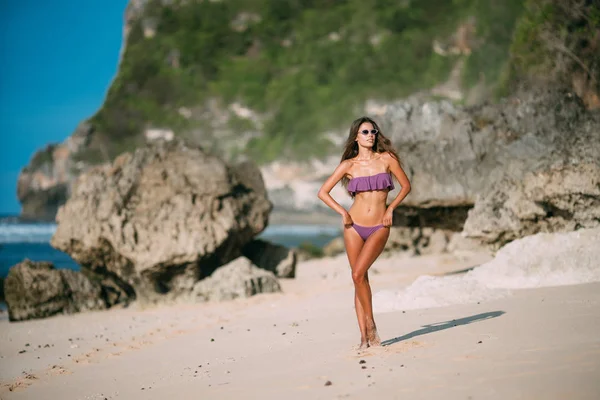 Sexy abbronzata ragazza in costume da bagno e occhiali da sole a riposo sulla spiaggia con sabbia e grandi pietre — Foto Stock