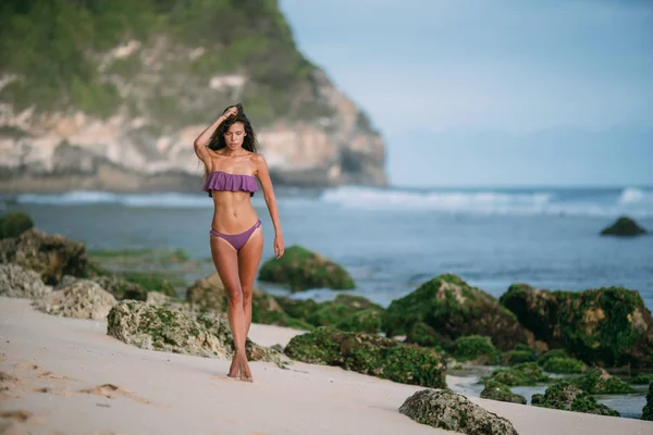 Сексуальная женщина в фиолетовом купальнике позирует на песчаном пляже с большими камнями на заднем плане — стоковое фото