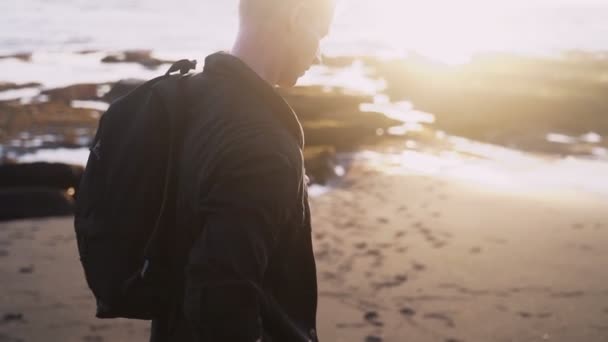 Guy a hátizsákban sétál végig fekete homokos strand napnyugtakor, lencse fáklyát. Steadicam lövés, lassított — Stock videók