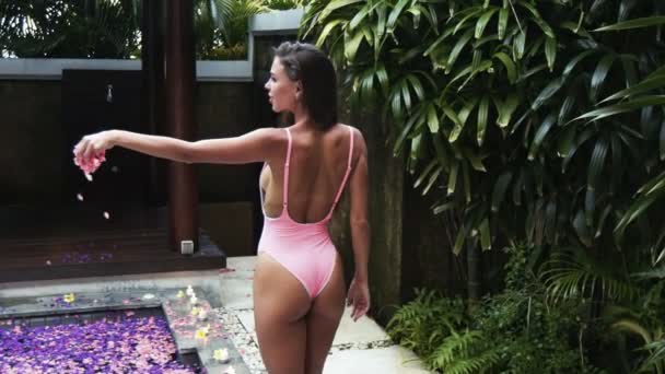 Backside view of sexy girl in pink swimsuit throws petals into pool full of flowers, slow motion — Stock Video