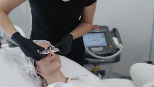 Las manos de cámara lenta del esteticista en guantes se pone gafas protectoras especiales en los ojos del paciente — Vídeo de stock