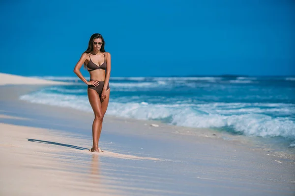 Sexy modello abbronzato con occhiali da sole in costume da bagno marrone in posa sulla spiaggia di sabbia bianca — Foto Stock