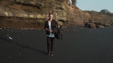 Sırt çantası olan adam siyah kum plaj boyunca yürür, akrobatik hile yapar. Yavaş çekim, steadicam çekim