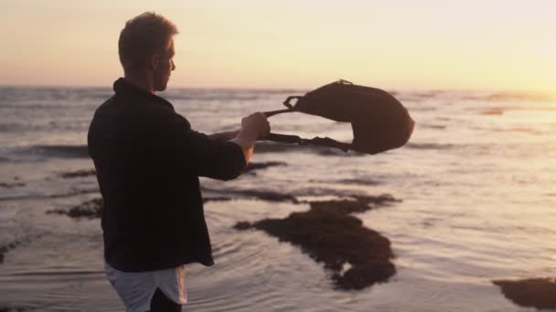 Young man in stylish modern clothes with backpack watching gold sunset at ocean beach. Slow motion — Stock Video