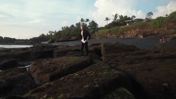 Man in stylish clothes with backpack walks on rocky black sand beach. Steadicam shot, slow motion — Stock Video