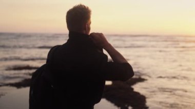 Sırt çantası okyanus sahilinde altın gün batımını izleyen adamın arka yüzü. Yavaş çekim, steadicam çekim