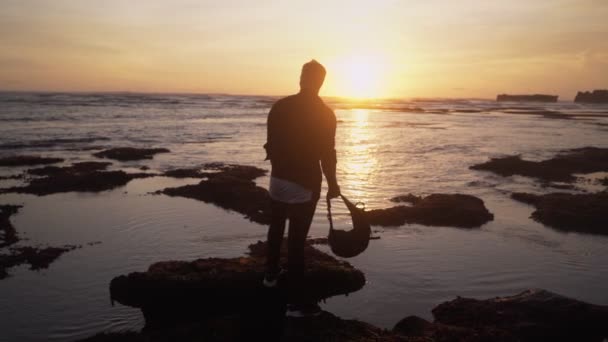 バックパックを持つ男のシルエットは、海のビーチに立って、美しい金の夕日に見えます。スローモーション — ストック動画