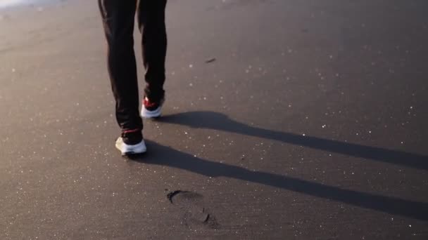 Cerrar las piernas del hombre en zapatillas de deporte elegantes caminando sobre arena brillante negro y deja huellas en la playa — Vídeos de Stock