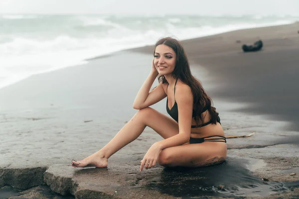 Porträtt av lycklig tjej med vackert leende i baddräkt poserar på svart sandstrand — Stockfoto