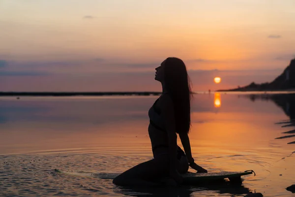 Silueta chica sexy en traje de baño acostado y posando en la tabla de surf en la playa durante la puesta del sol — Foto de Stock