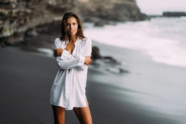 Joven chica sexy en camisa blanca y traje de baño descansando en la playa de arena negra . —  Fotos de Stock