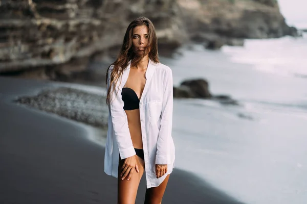 Young sexy girl in white shirt and swimsuit resting on black sand beach. — Stock Photo, Image