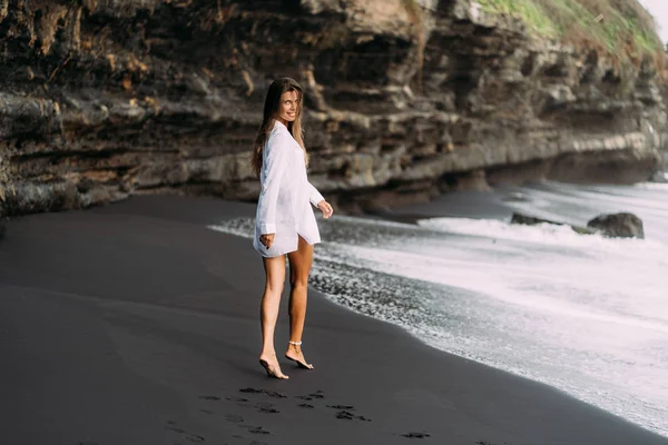 Joven chica sexy en camisa blanca y traje de baño descansando en la playa de arena negra . — Foto de Stock