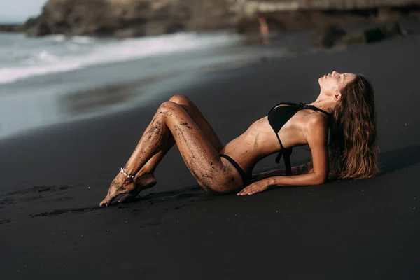 Attractive sexy girl in swimwear rests on black volcanic sand beach. — Stock Photo, Image